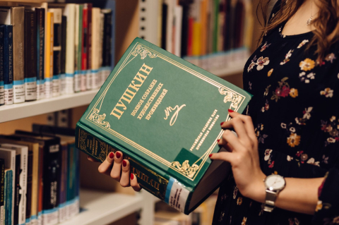 Девушка держит томик Пушкина в зелёном переплёте у книжных полок библиотеки 