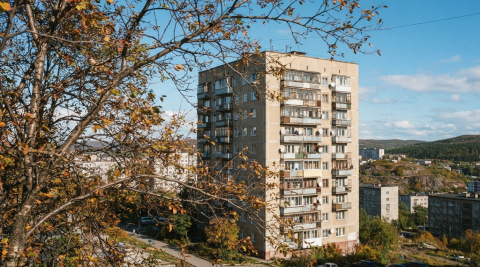 Панельная многоэтажка с застеклёнными балконами за осенними ветками на фоне синего неба 