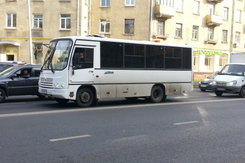 Белый небольшой автобус едет по городской улице мимо жилого дома 