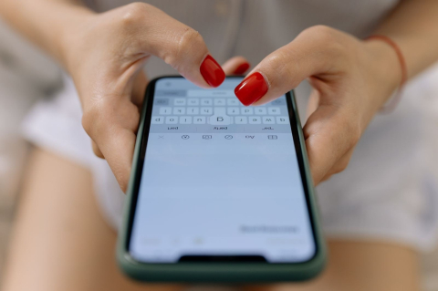 Пальцы с красным маникюром печатают сообщение на смартфоне в зелёном чехле 