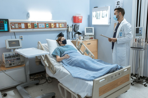 Пациент в маске лежит на больничной койке, рядом врач в белом халате с планшетом 
